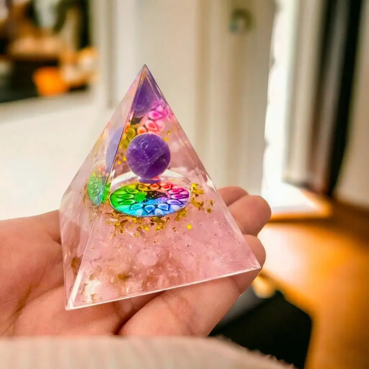 Pessoa segurando uma pirâmide de Orgonite com Quartzo Rosa, com a representação dos 7 Chakras e uma esfera central, evidenciando a beleza e o detalhe do produto.